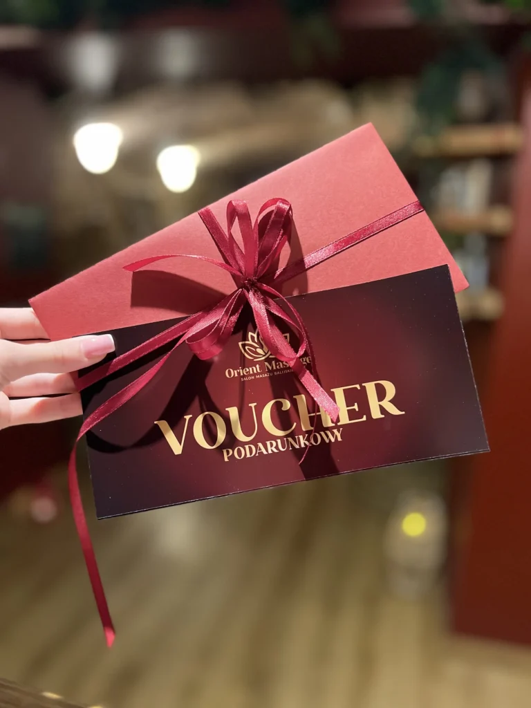 Voucher podarunkowy z czerwoną kopertą i kokardą trzymany w dłoni.