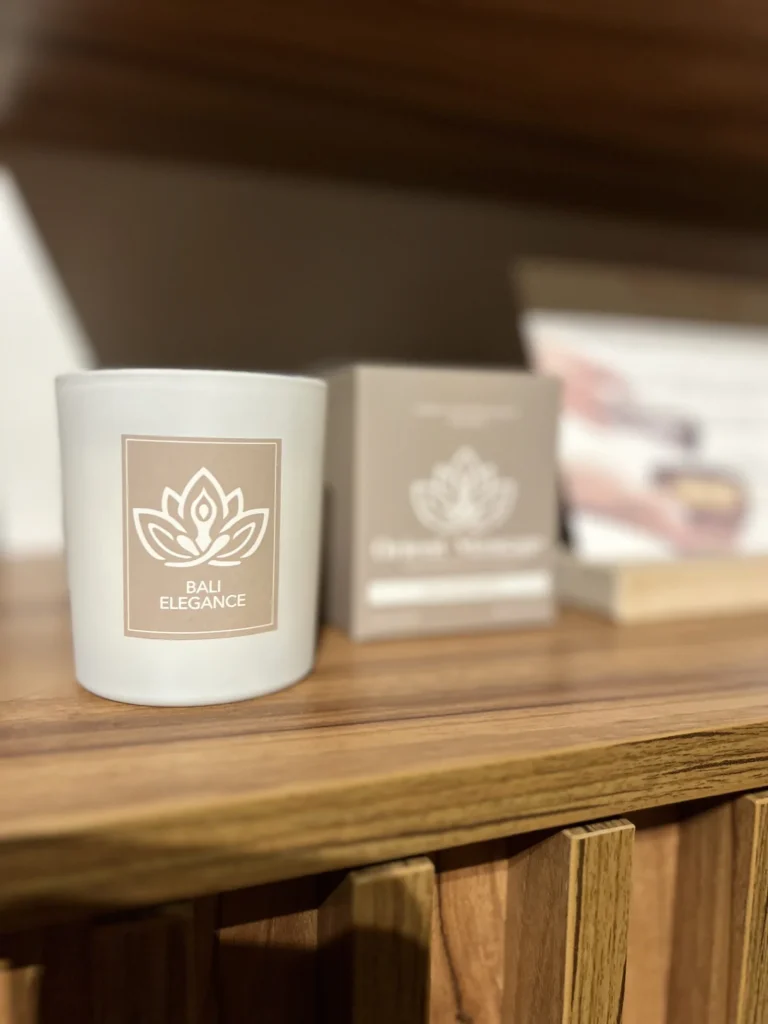 Biała świeca Bali Elegance z logo Orient Massage na drewnianej półce.