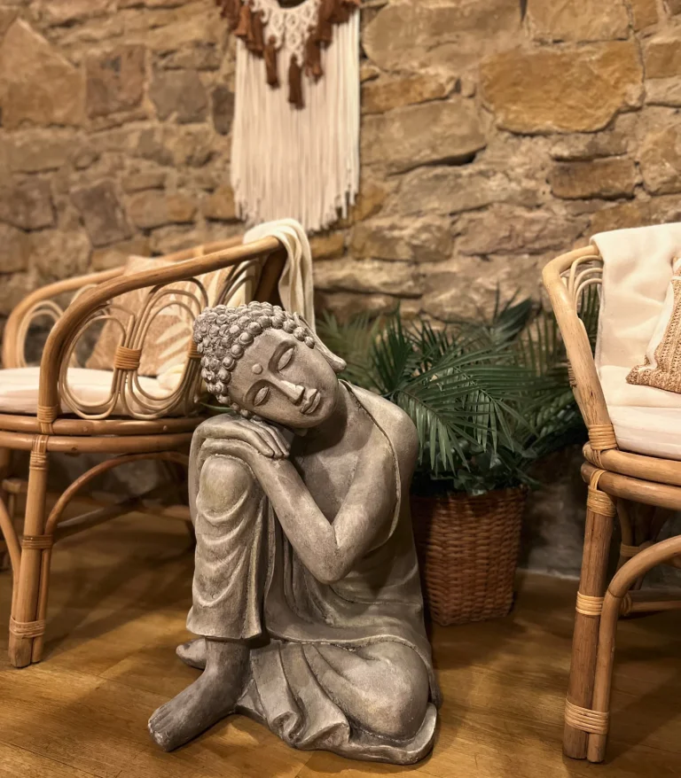 Figura siedzącego Buddy między rattanowymi fotelami przy kamiennej ścianie.
